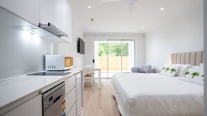 Une chambre d’hôtel moderne dotée d’une kitchenette, d’un lit avec une literie blanche et des coussins à motifs de palmiers, d’un téléviseur à écran plat et d’une grande fenêtre offrant une vue sur la verdure.