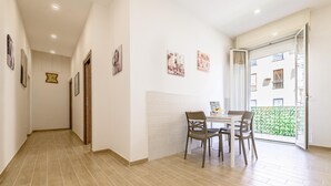 Um corredor com área de jantar, apresentando uma mesa com cadeiras, um relógio na parede e uma porta que dá para uma varanda com vista para edifícios.