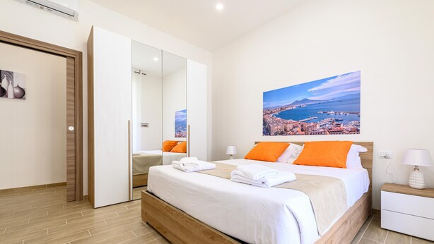 A modern bedroom with a wooden bed, white bedding, and orange pillows. A large mirror and a nightstand with a lamp are visible. - Domus Romeo - Camera 1 (Naples)