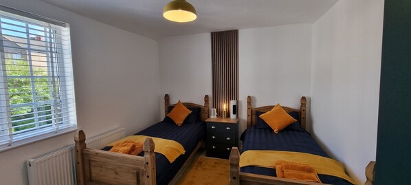 A bedroom with two wooden beds, each with a blue mattress and an orange blanket. There is a nightstand with a lamp and a radio between the beds. A window with blinds is on the left, and a radiator is below the window. - The Hideaway ll /enclosed Garden/quiet/central (Southport)