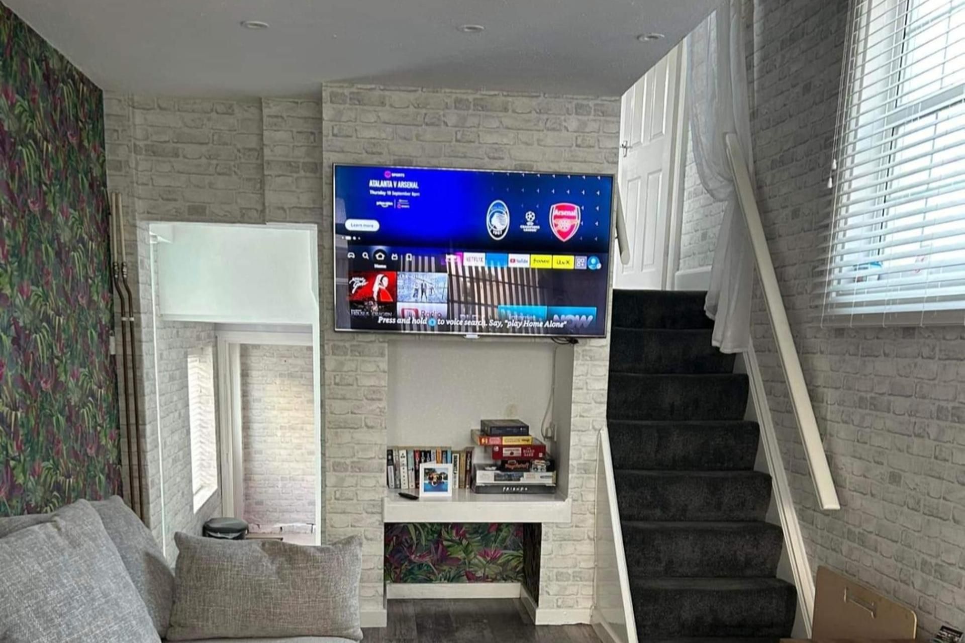 A living room with a flat-screen TV mounted on a brick wall, a staircase to the right, and a bookshelf with books and games below.