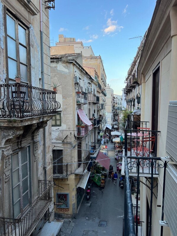 A narrow street with buildings on either side, balconies with railings, and a market area with stalls.