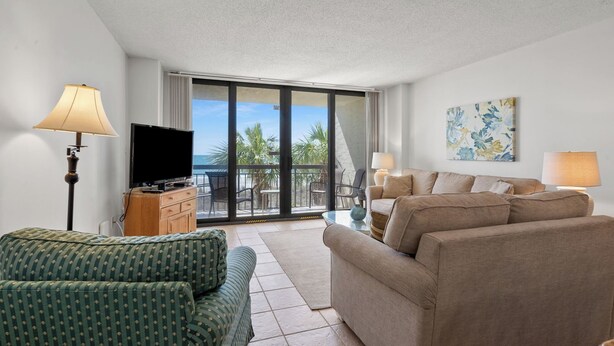A living room with a sofa, a TV, and a balcony with a view of palm trees. - ShorehamTowers Oceanfront Resort (NORTH MYRTLE BEACH)
