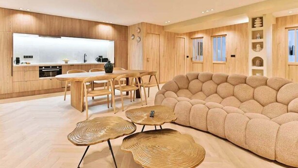 Eine moderne Küche mit Holzausstattung, ein Essbereich mit rundem Tisch und Stühlen, ein beiges Sofa und ein dekoratives Wandregal.
