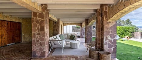 A covered patio area with stone pillars, a sofa set, and a view of a green lawn.