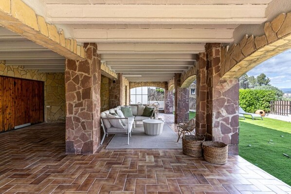 A covered patio area with stone pillars, a sofa set, and a view of a green lawn. - Renovated villa, pool, barbecue, and just 15 minutes from Barcelona. (Sant Cugat del Vallès)
