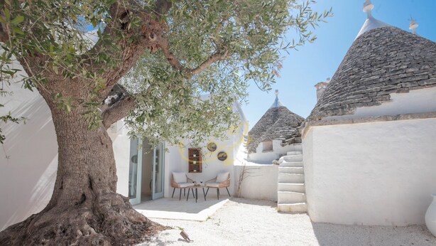 Un edificio tradicional de paredes blancas y techo de piedra, con un gran árbol en primer plano y una pequeña zona de estar con dos sillas.