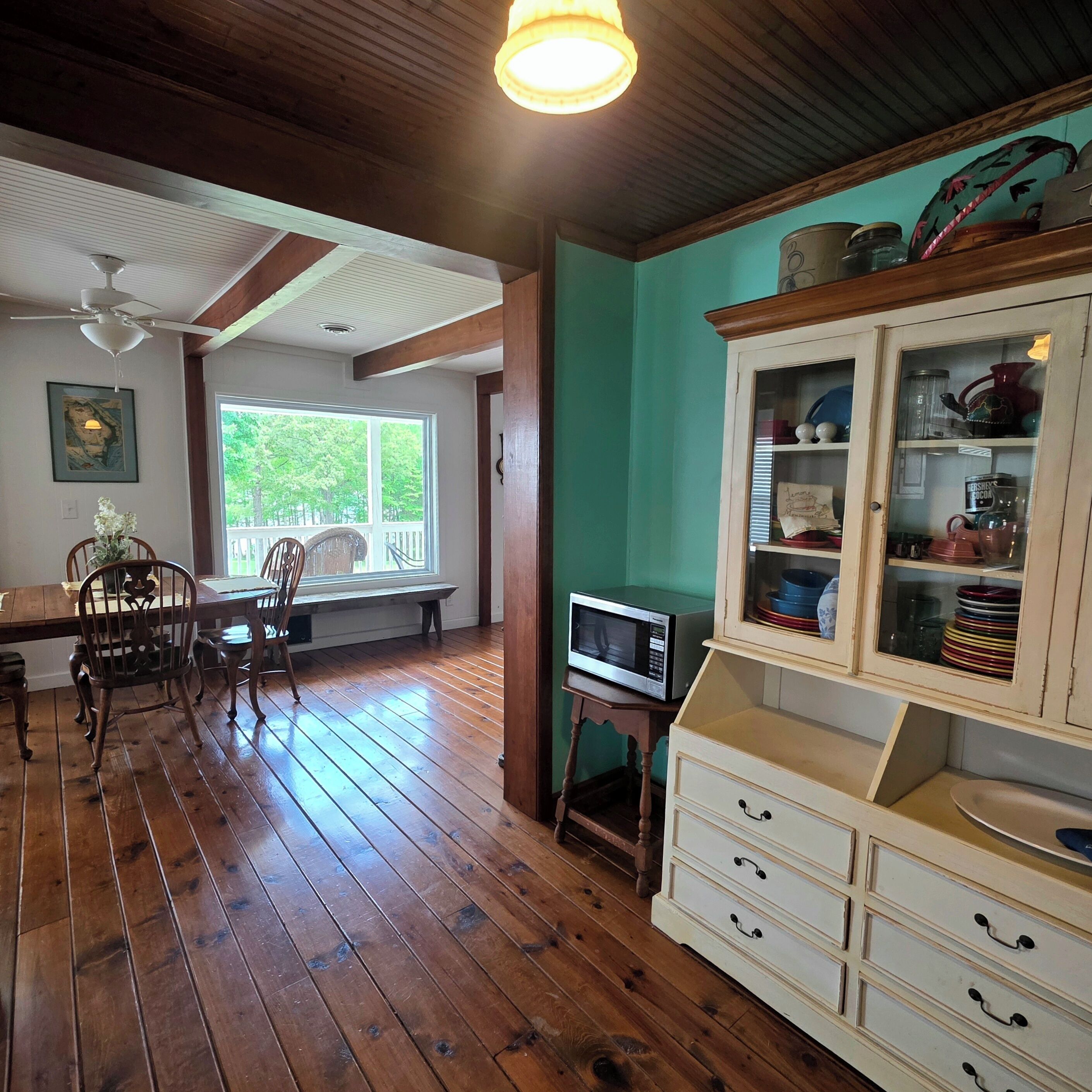 A dining area with wooden flooring, a table, chairs, a ceiling fan, and a window with a view of trees.