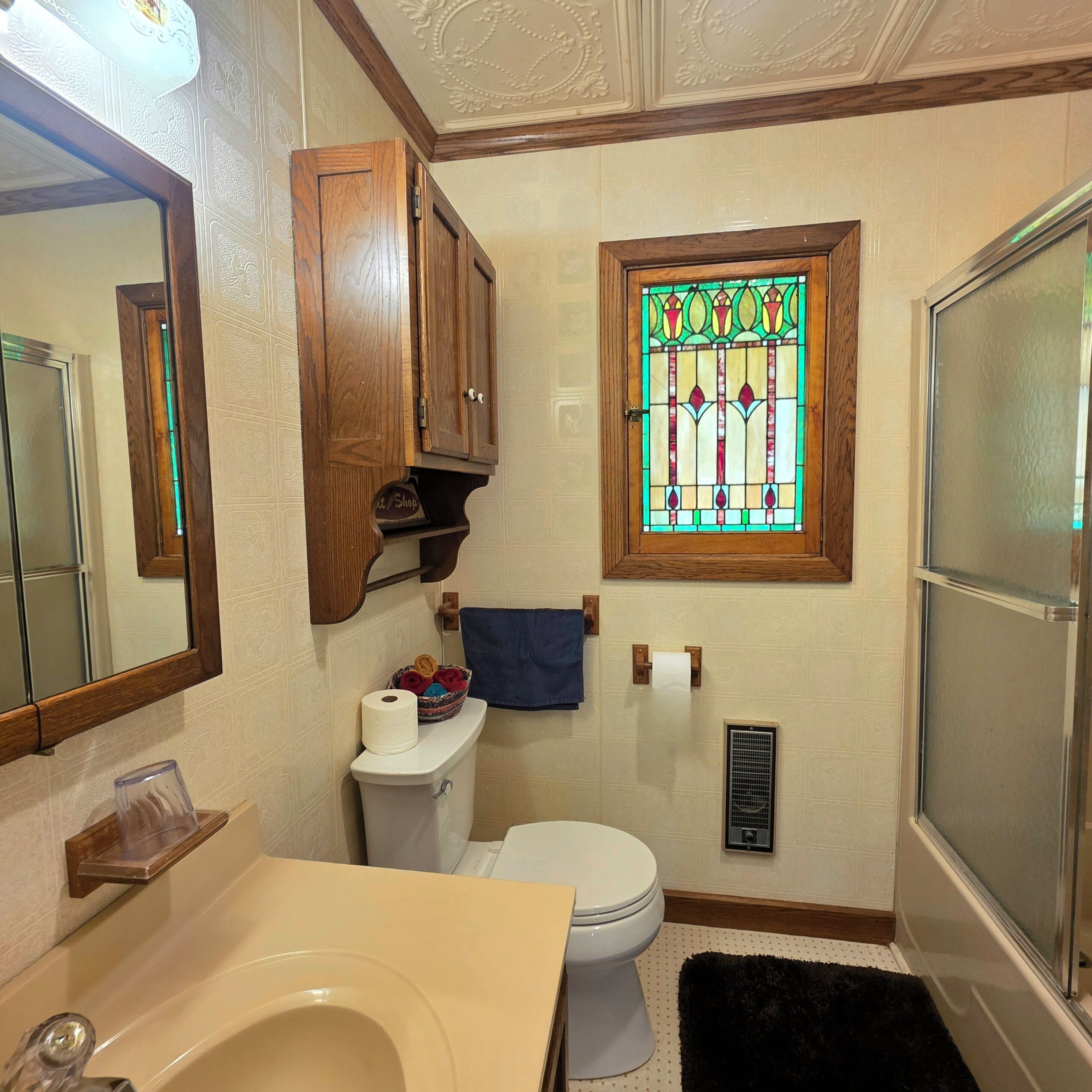 A bathroom with a toilet, a sink, a mirror, a wooden cabinet, a stained glass window, and a shower area.