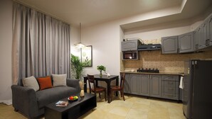 A modern living room with a sofa, coffee table, and dining table. A kitchen with grey cabinets and appliances is visible. - BedChambers Serviced Apartments SushantLok (Gurugram)