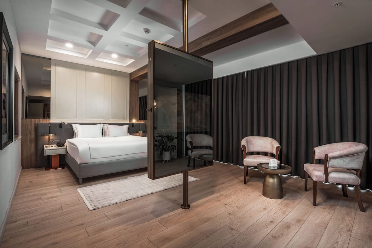 A modern bedroom with a large bed, bedside tables, a mirror, and two armchairs.