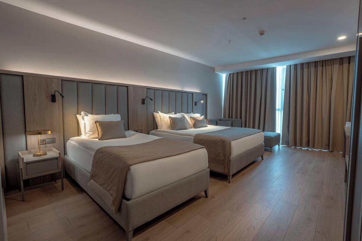 A modern hotel room with two beds, a nightstand, a lamp, and a large window with curtains.