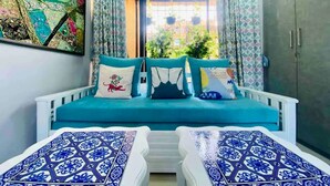 A cozy room with a blue sofa bed, patterned cushions, and a view of greenery through a window. - Charming 3 Bed 2 Bath in Bandra West with Balcony
 (Mumbai)