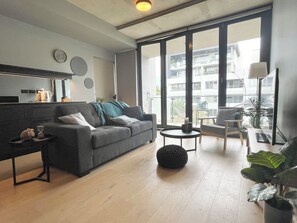 Uma sala de estar moderna com um sofá cinza, uma mesa de centro e um banquinho redondo preto. Há uma janela grande com vista para uma varanda e edifícios.