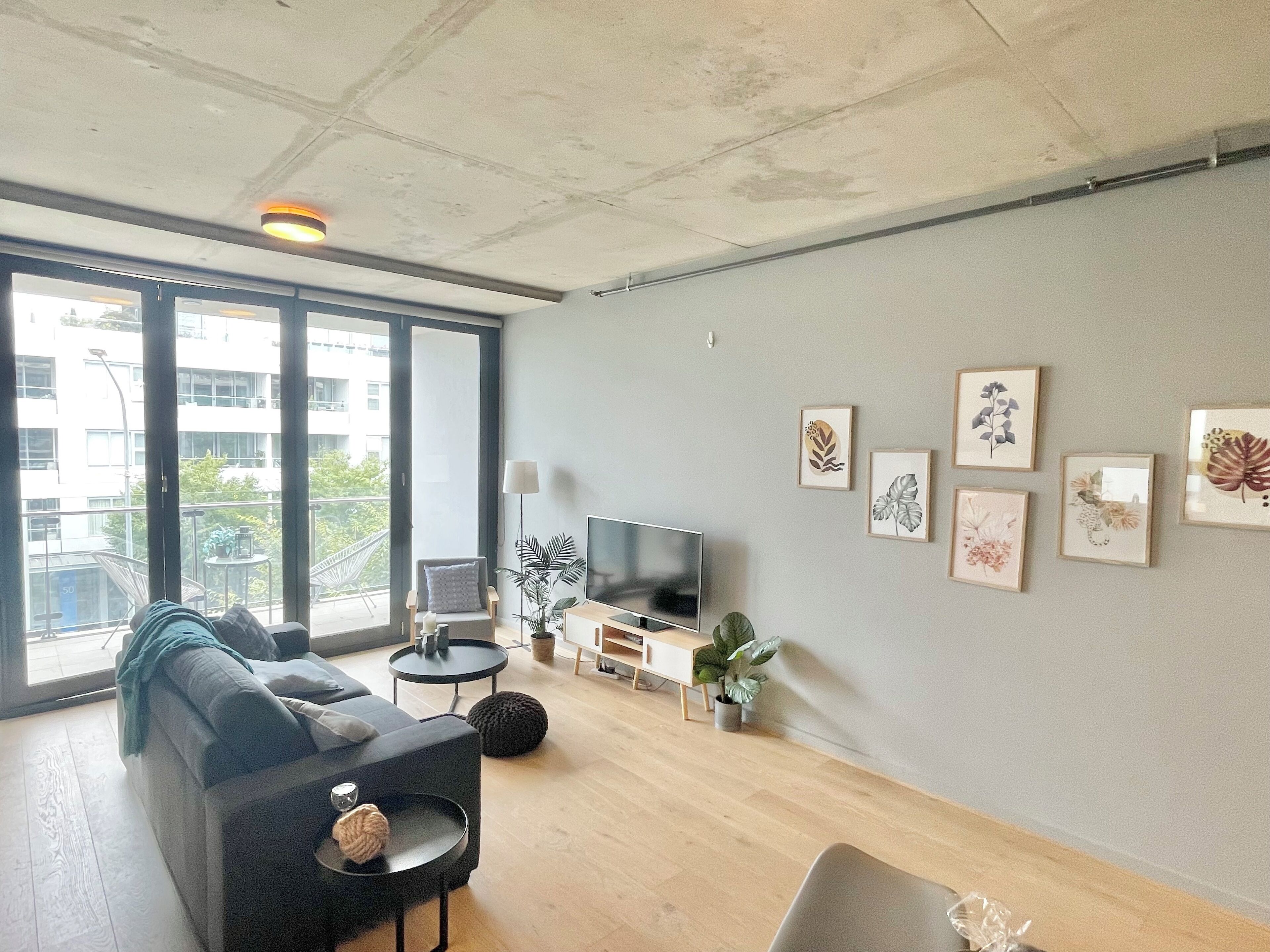Un salon moderne avec un mur gris, une grande fenêtre, un canapé gris, un téléviseur et plusieurs œuvres d’art encadrées.