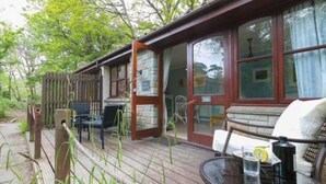 Una terraza de madera con mobiliario exterior, un muro de piedra y un ventanal que da a una habitación.
