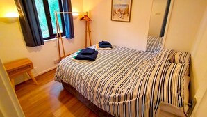 Un dormitorio con colcha a rayas, una lámpara de pie, una ventana con cortinas, un cuadro enmarcado y un espejo.