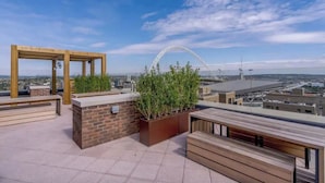Una terrazza sul tetto con panche in legno, un fioriere in mattoni con piante verdi e vista su un paesaggio urbano che include una ruota panoramica.