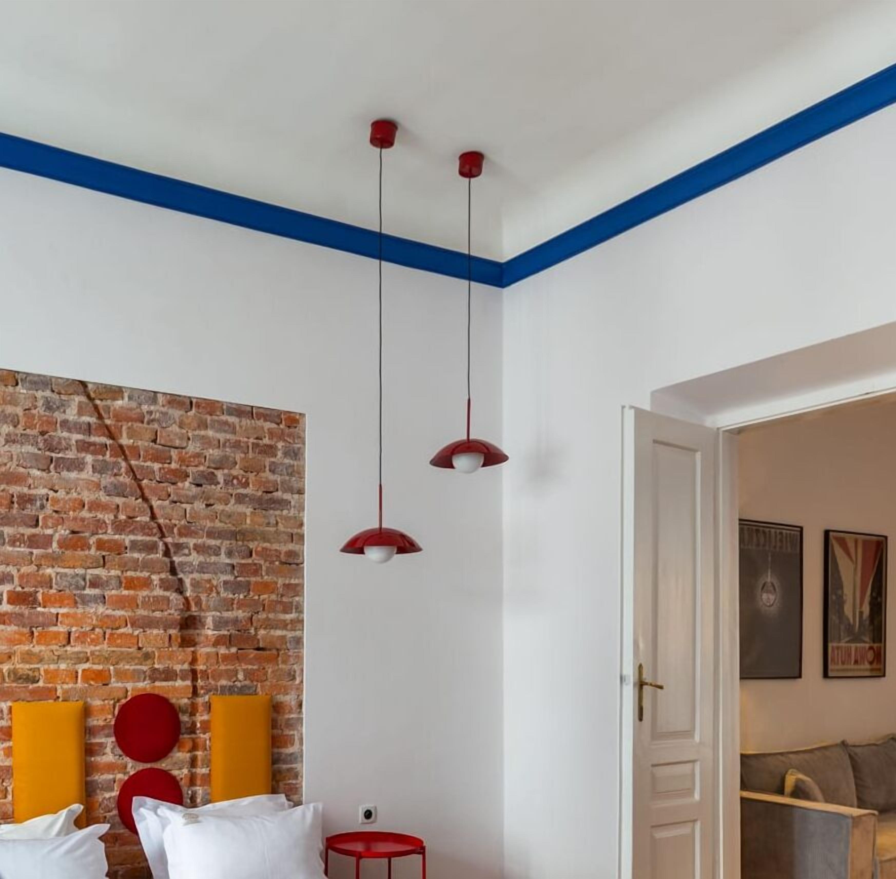 Una habitación con pared de ladrillo, techo blanco con detalles azules, cama con almohadas blancas y rojas, una mesita de noche roja y una puerta que da a otra habitación.