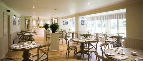 A spacious dining area with wooden flooring, round tables, and wicker chairs.