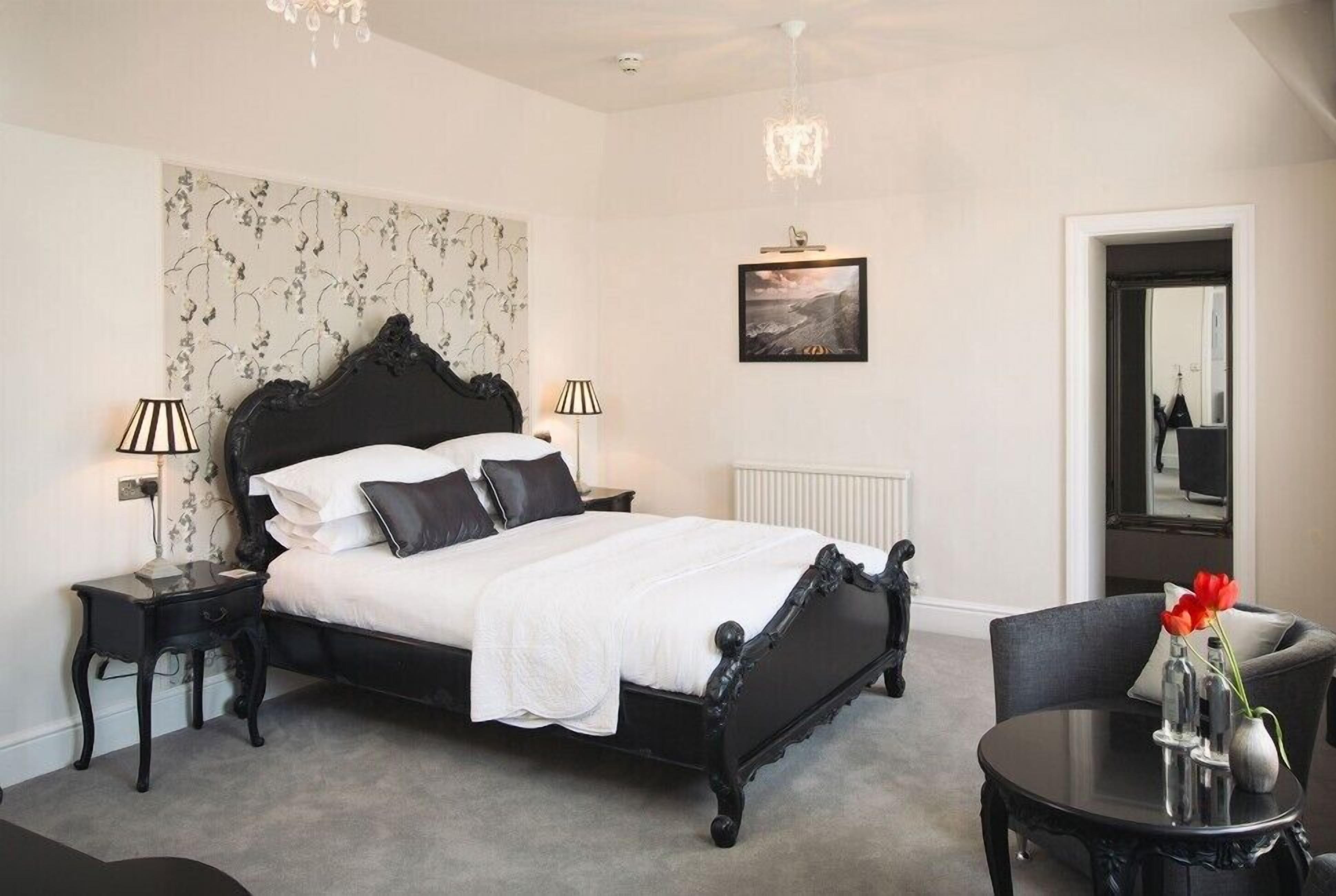 A bedroom with a black ornate bed, two bedside tables with lamps, a round table with a vase, and a framed picture on the wall.