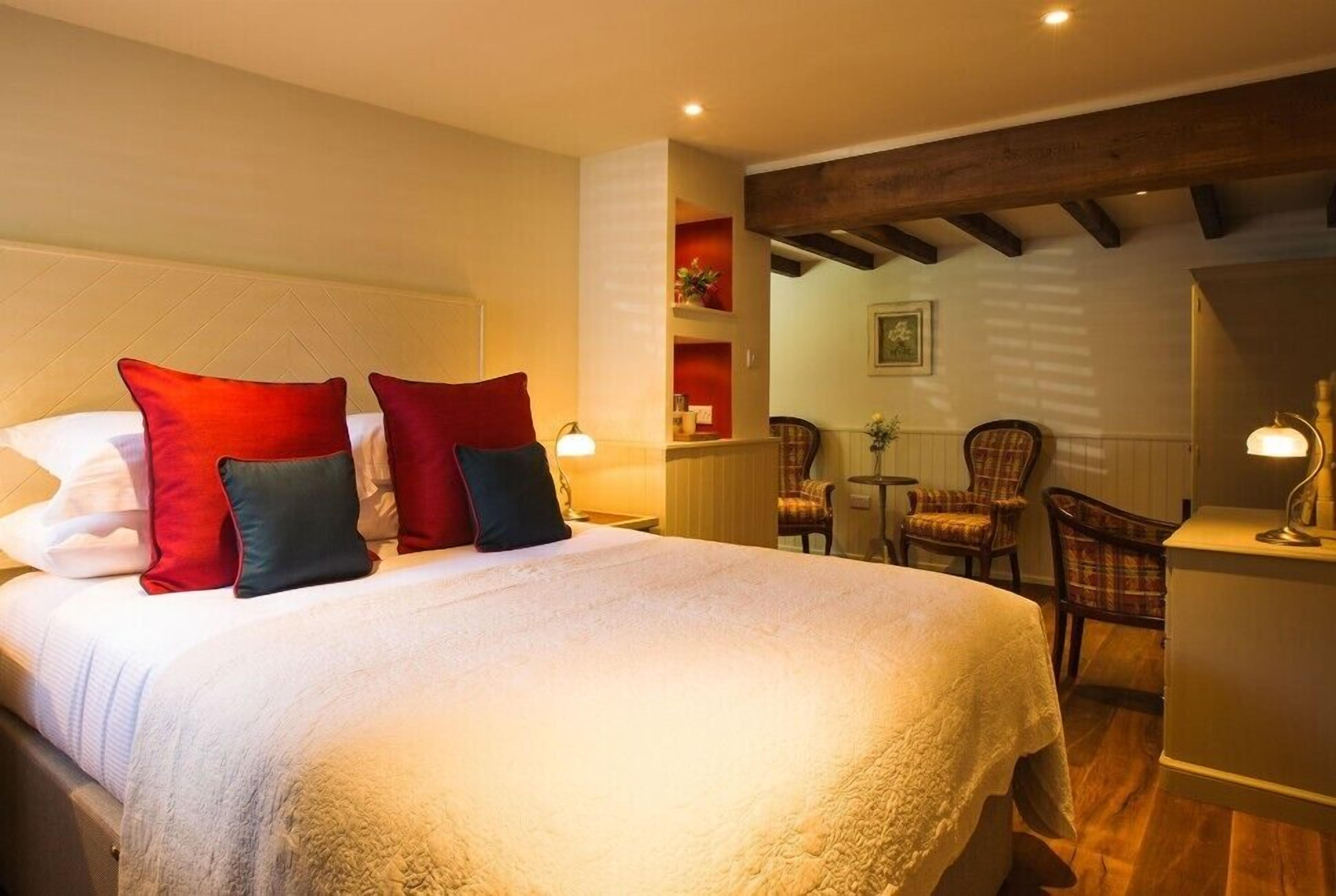 A hotel room with a large bed, red and black pillows, a wooden ceiling, and a seating area with chairs and a table.