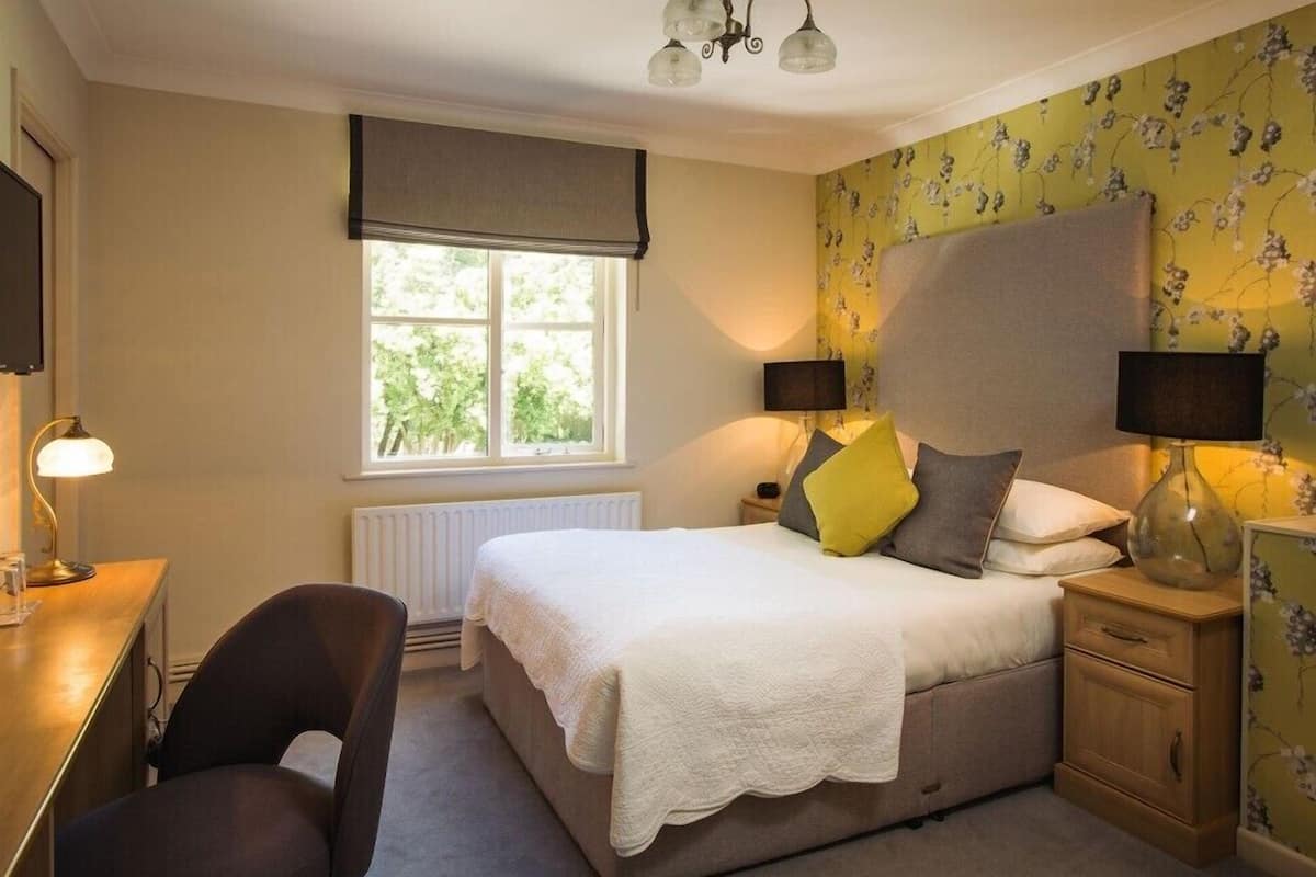 A bedroom with a bed, bedside lamps, a chair, a desk, and a window with a view of greenery.