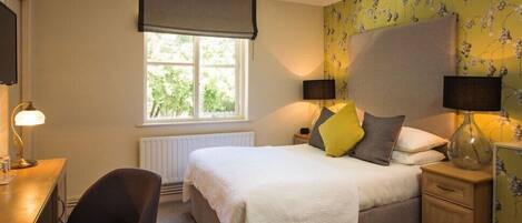 A bedroom with a bed, bedside lamps, a chair, a desk, and a window with a view of greenery.