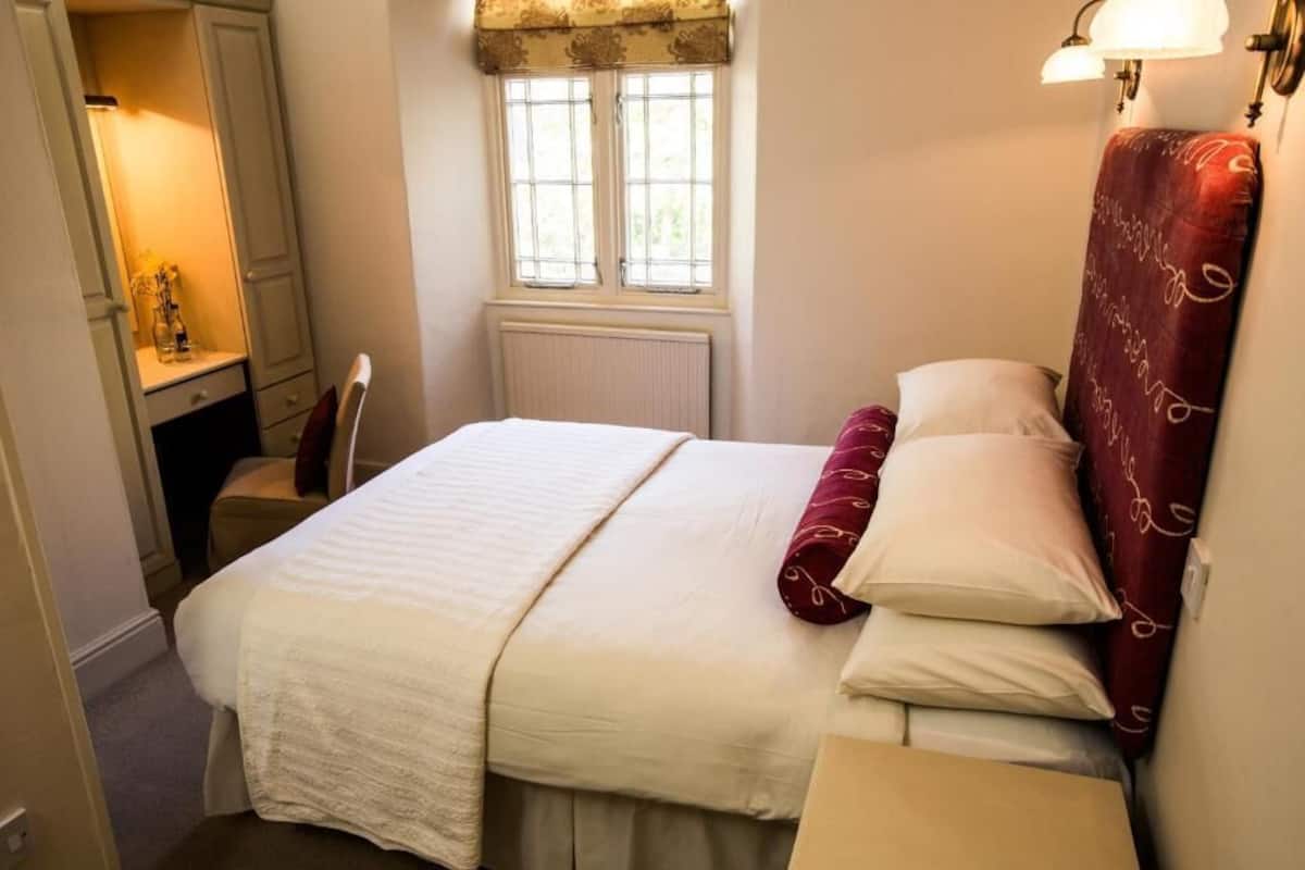 A neatly arranged bedroom with a bed, a bedside table, a window with curtains, and a wall-mounted lamp.