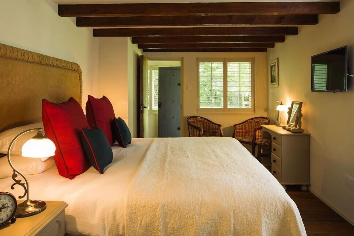 A bedroom with a bed, a nightstand with a lamp, a chair, a desk with a lamp, and a window with blinds.