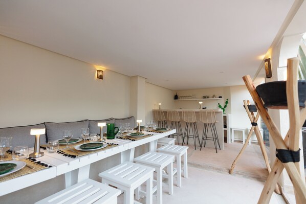 A modern kitchen with a long dining table set for six, white bar stools, and a bar area with high stools.