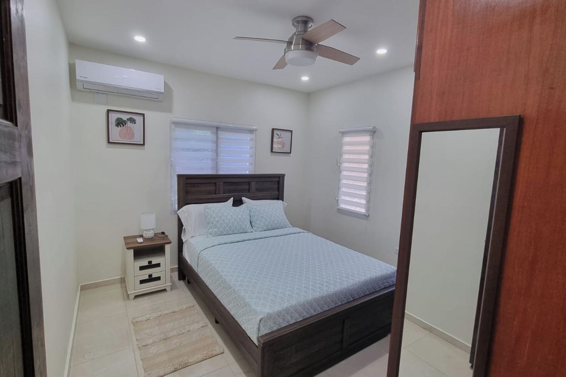 Un dormitorio con cama, mesita de noche, aire acondicionado instalado en la pared, ventilador de techo y dos ventanas con persianas.
