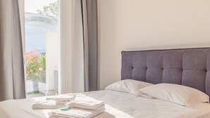 A neatly made bed with white linens and a grey headboard, a window with curtains, and a view of greenery outside. - Rosa dei Venti Ovest (Padenghe sul Garda)