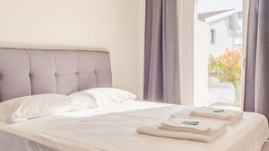 A neatly made bed with white linens and pillows, a grey upholstered headboard, and a window with grey curtains. - Rosa dei Venti Est in Padenghe sul Garda (Padenghe sul Garda)