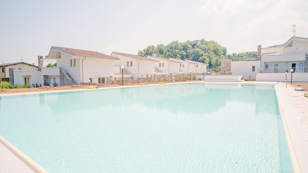 A swimming pool surrounded by modern buildings with a clear view of a forested hill in the background. - Rosa dei Venti Est in Padenghe sul Garda (Padenghe sul Garda)