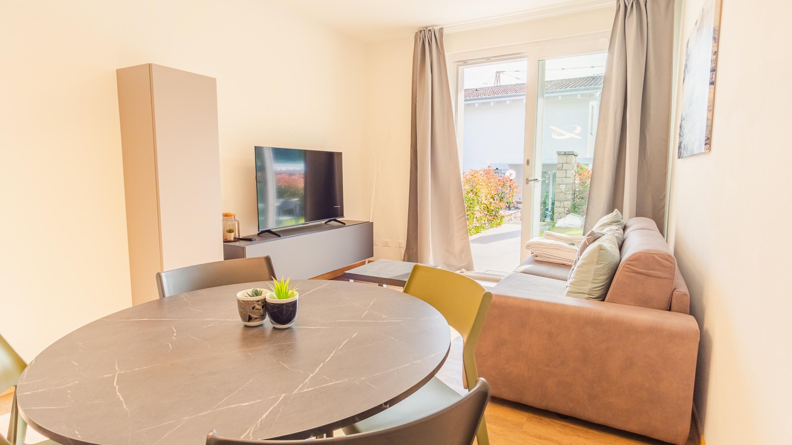 A modern living room with a round dining table, a sofa, a TV, and a balcony with curtains.