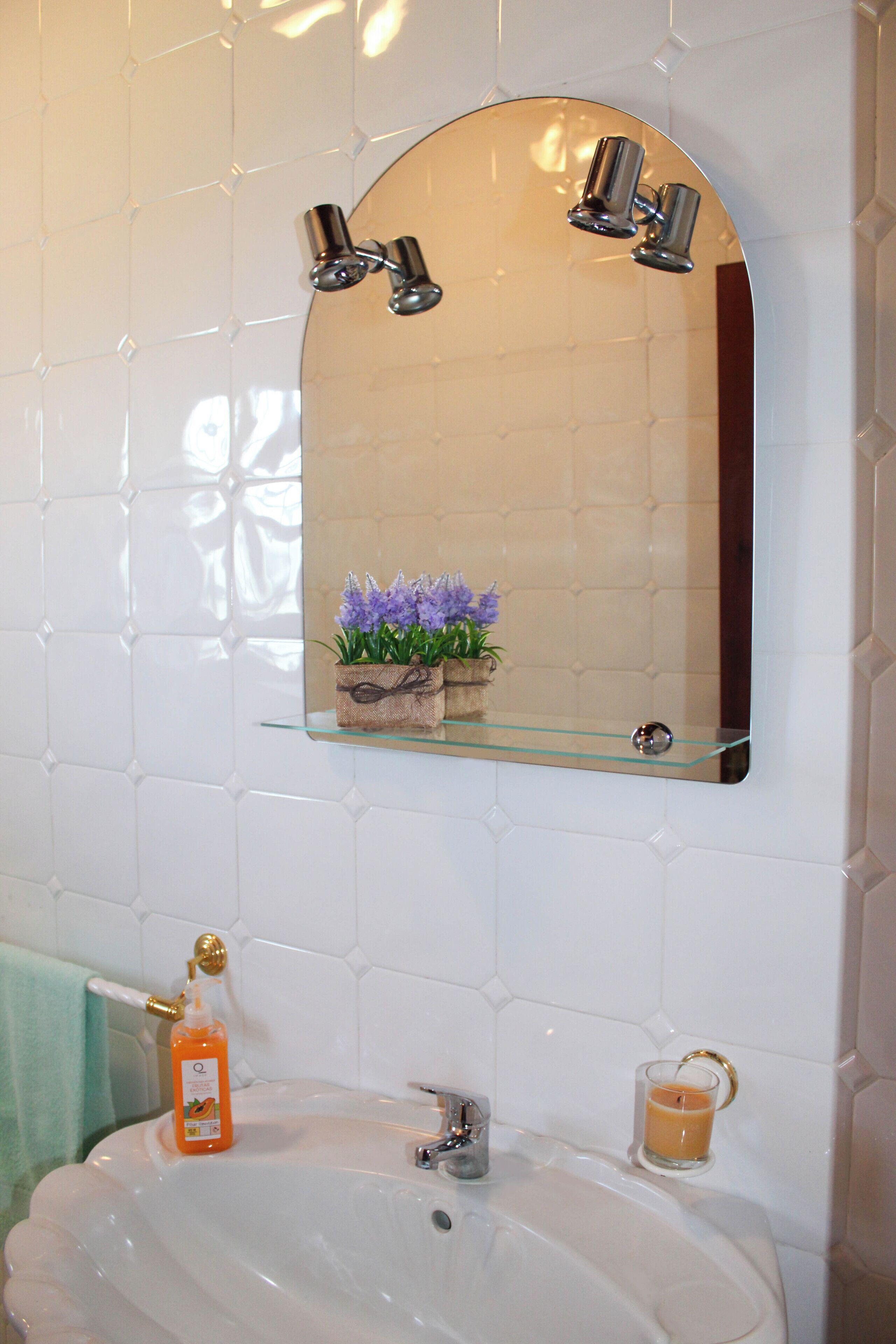 A bathroom with a white tiled wall, a glass shelf with a plant, a sink with a bottle of soap, and a glass of orange juice.