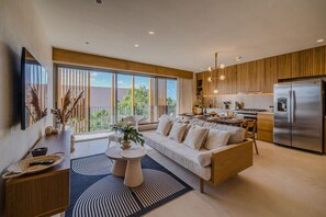 A modern kitchen with a built-in oven and refrigerator, a dining area with a sofa and coffee table, and large windows offering a view of greenery.