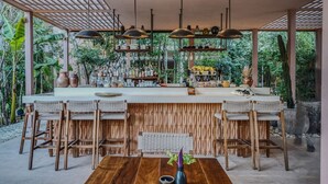 A bar area with wooden chairs and a long counter, surrounded by greenery and plants.