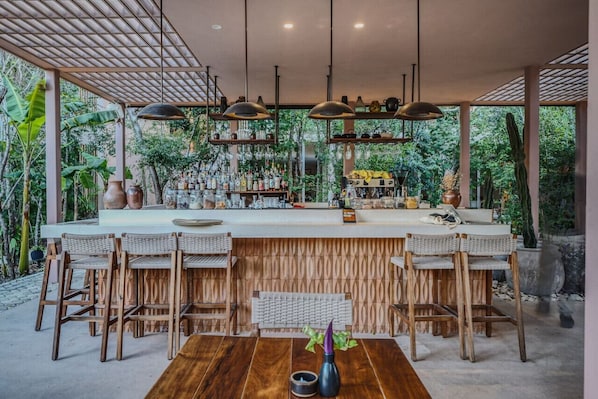 A bar area with wooden chairs and a long counter, surrounded by greenery and plants.