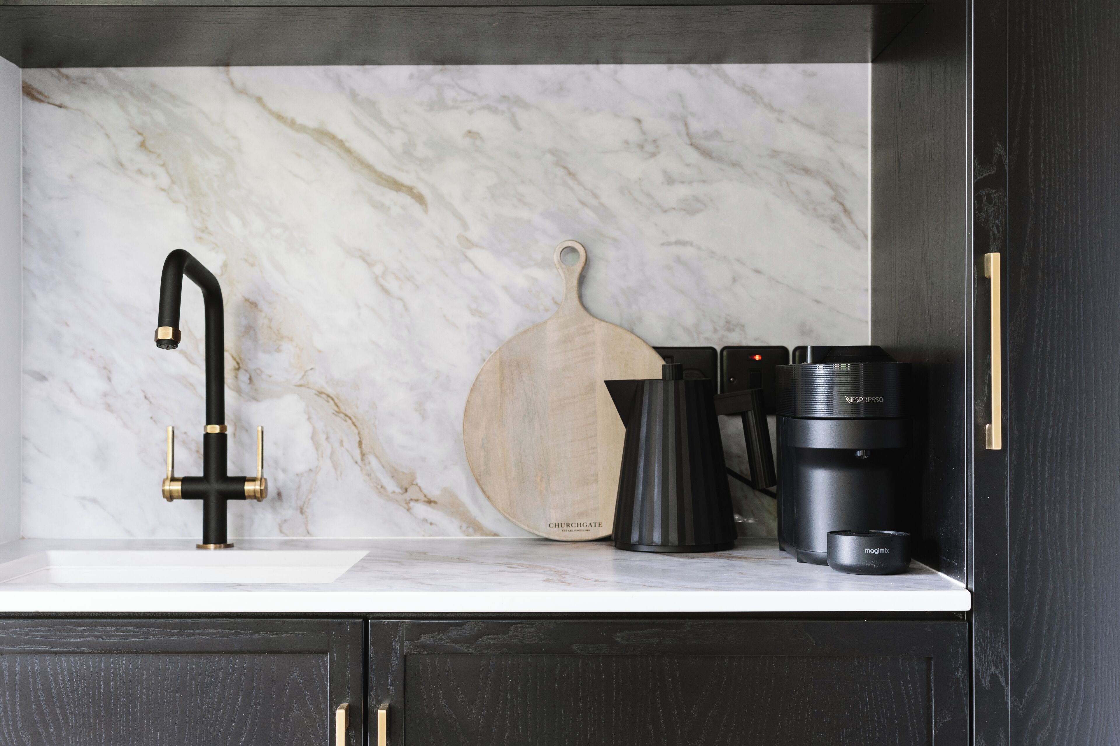 Una cocina con salpicadero de mármol, grifo negro, tabla de cortar de madera, cafetera y jarra de café.