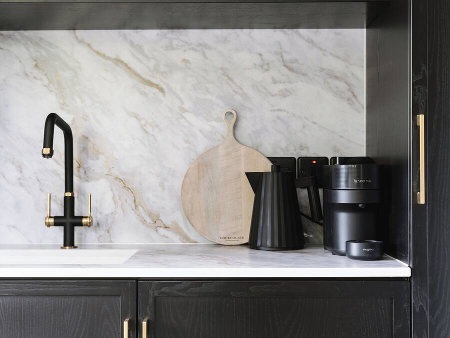Una cocina con salpicadero de mármol, grifo negro, tabla de cortar de madera, cafetera y jarra de café.