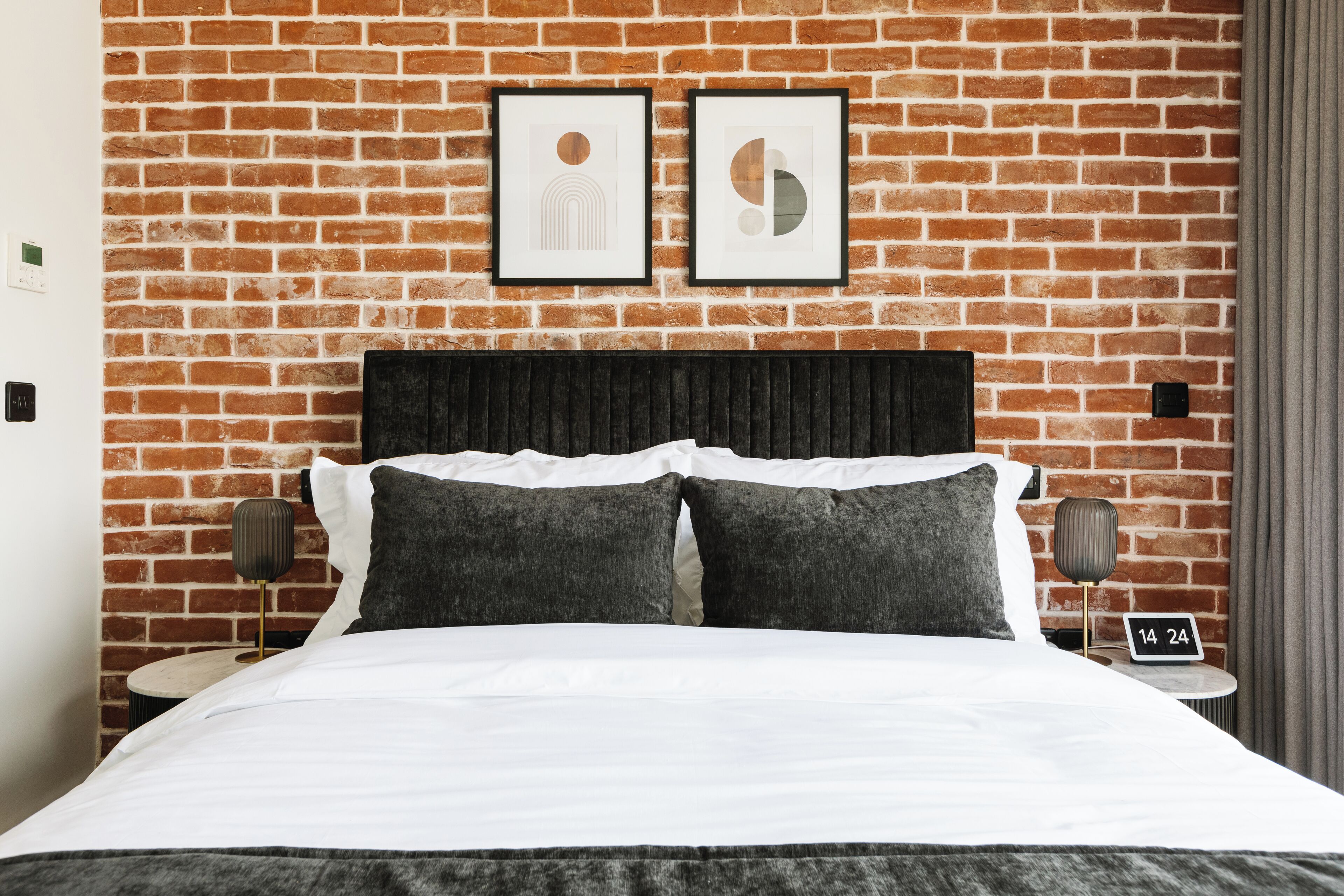 Quarto de hotel com parede de tijolos, cama com roupa de cama branca, duas almofadas, mesa de cabeceira com abajur e duas molduras com imagens na parede.