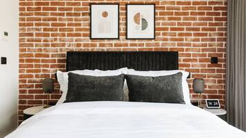Quarto de hotel com parede de tijolos, cama com roupa de cama branca, duas almofadas, mesa de cabeceira com abajur e duas molduras com imagens na parede.