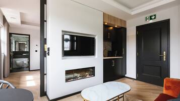 Une chambre d’hôtel moderne dotée d’un téléviseur à écran plat fixé au mur, d’une petite cheminée, d’un coin repas avec une table et des chaises, et d’une porte noire avec un panneau vert indiquant la sortie.
