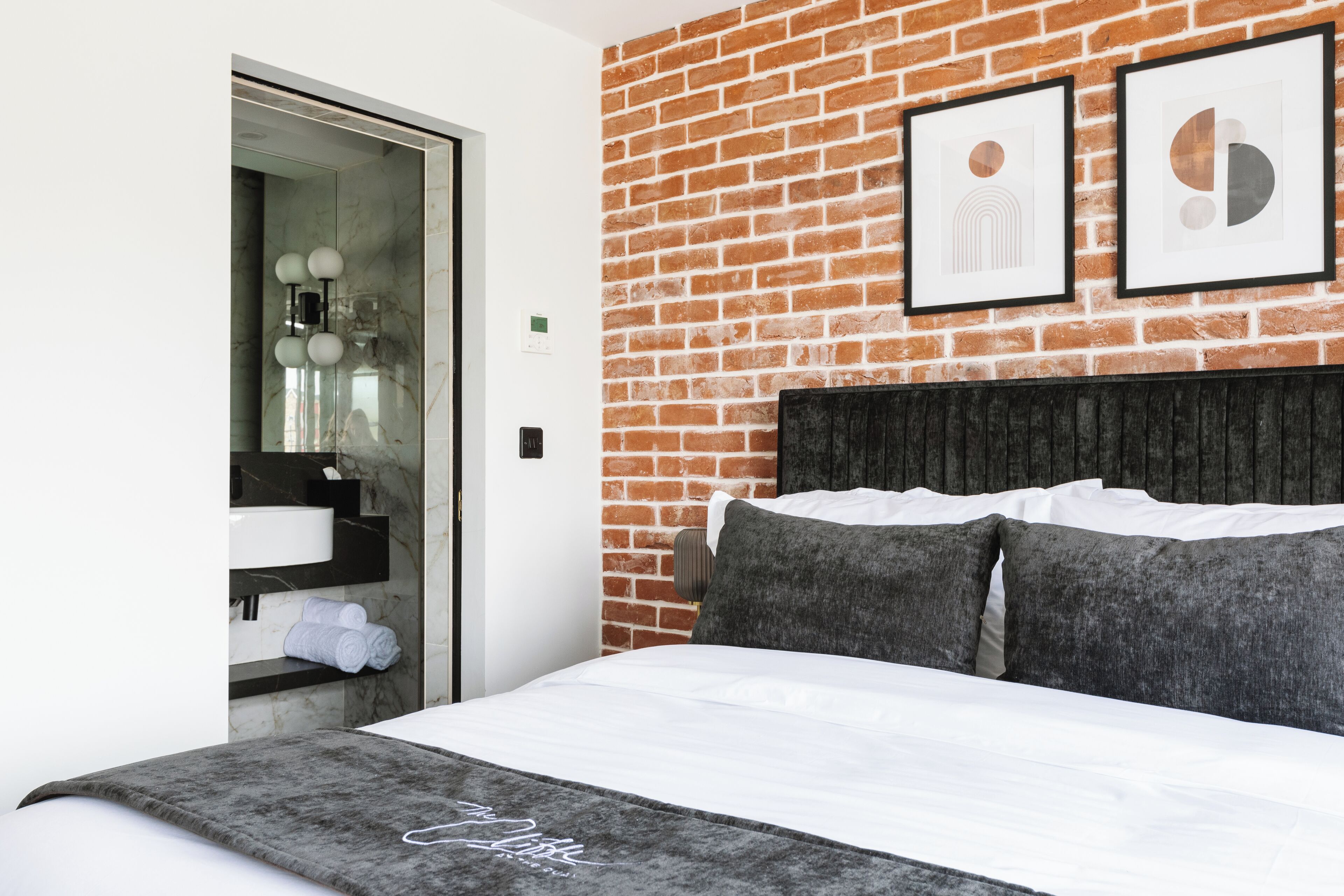 A modern hotel room with a brick wall, a bed with white linens, and two framed artworks above the bed.