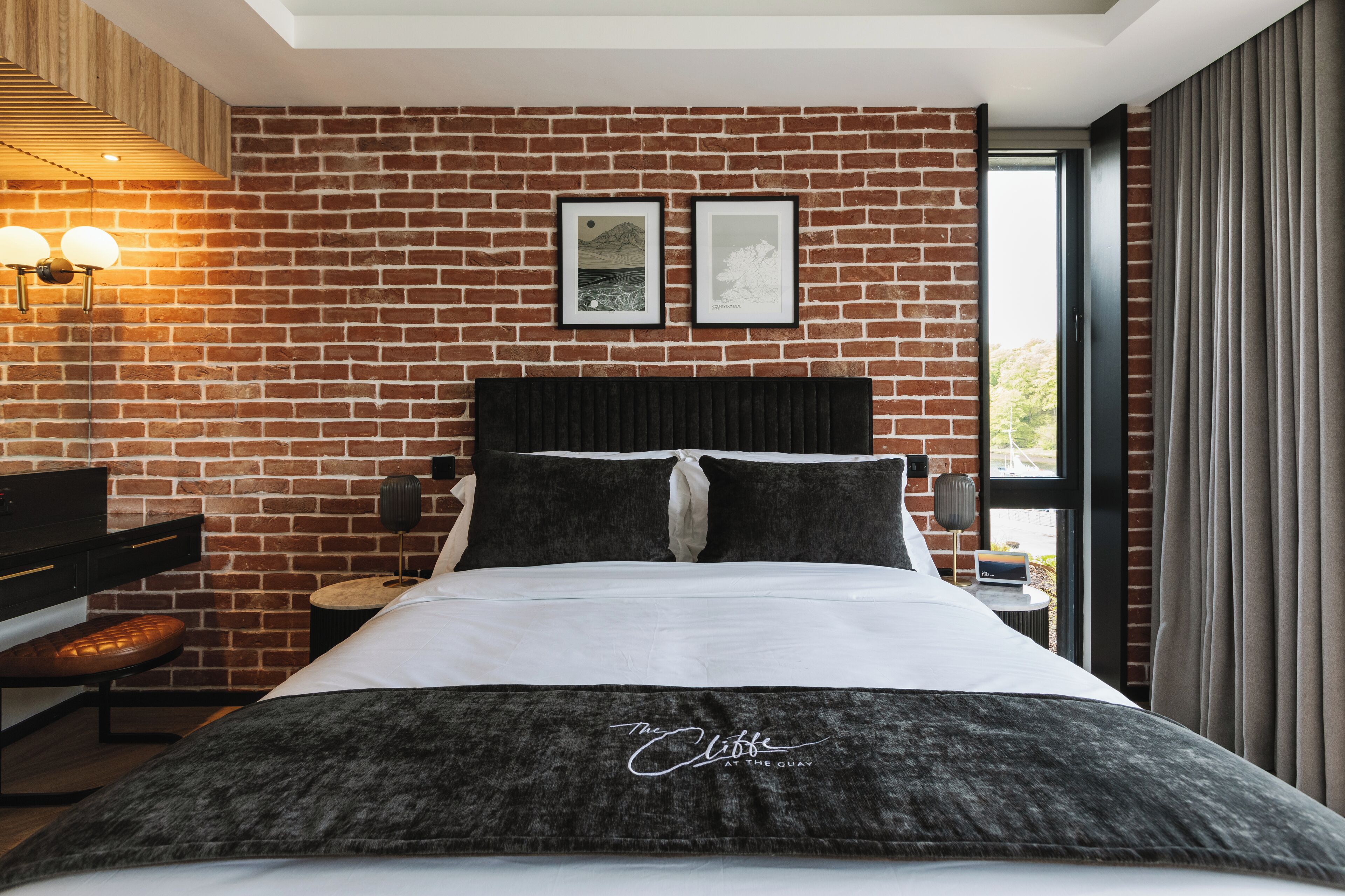 A hotel room with a brick wall, a bed with a black headboard, two framed pictures on the wall, a desk with a lamp, and a window with curtains.
