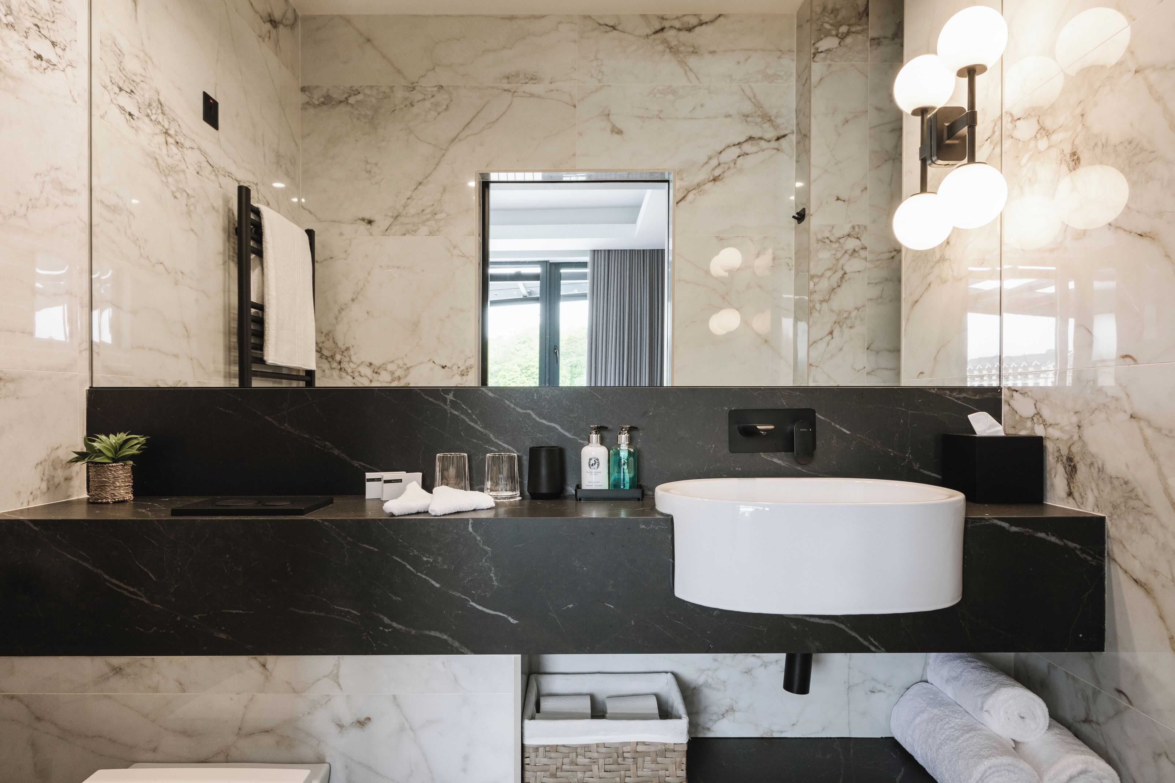 A modern bathroom with a marble wall, a black countertop, a white sink, and a mirror with lights.