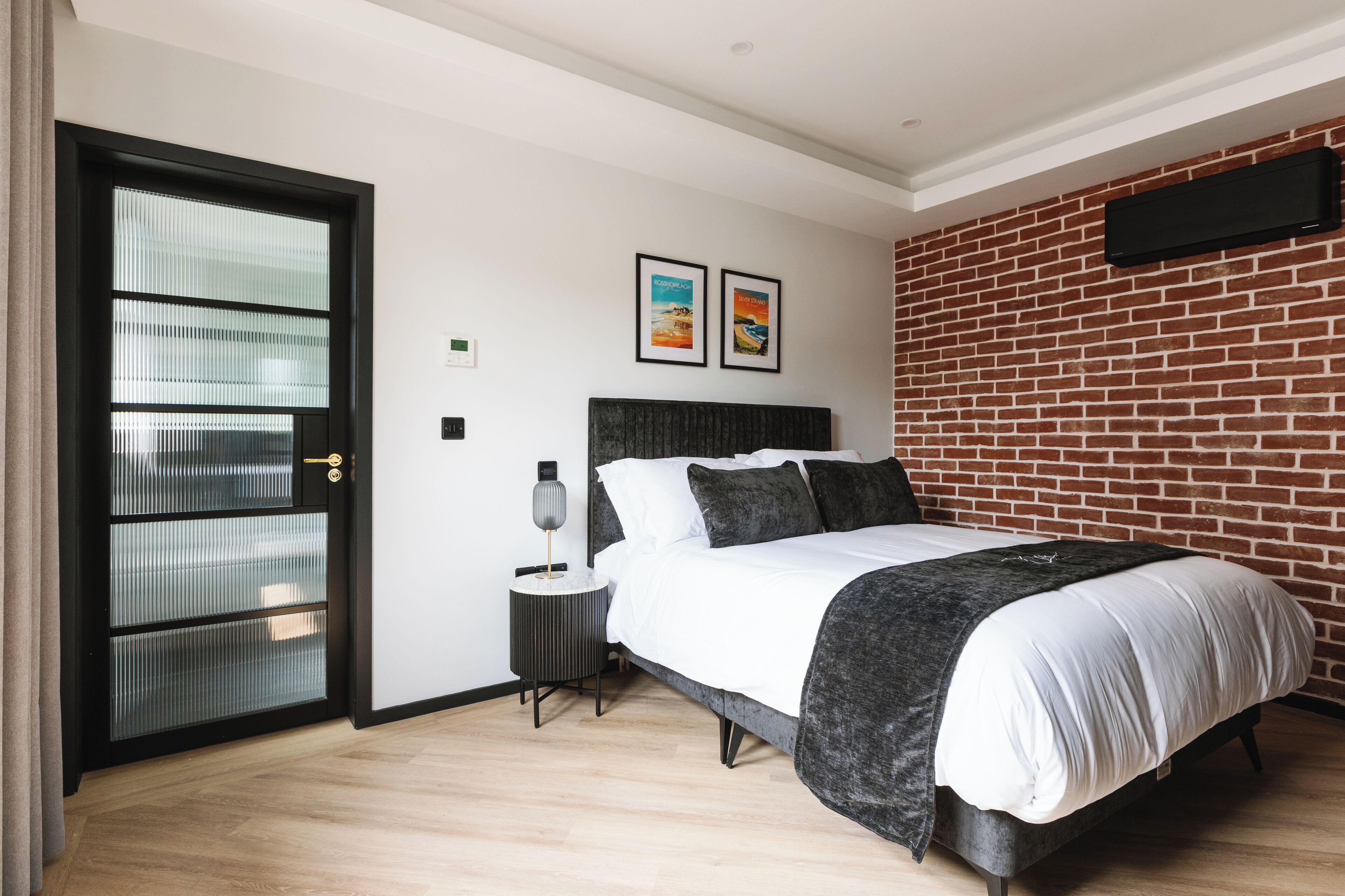 A modern bedroom with a brick-patterned wall, a bed with a dark headboard, and a side table with a lamp.