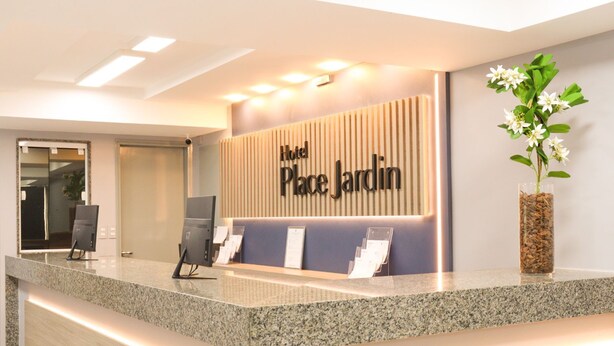 A hotel reception desk with a sign reading "Hotel Place Jardin" and a potted plant on the right. - Hotel Place Jardin (Garanhuns)